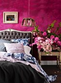 Double bed with button-tufted headboard and white, blue and grey bed linen next to bouquet on bedside table against purple-painted wall