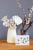 White-painted artificial flowers in simple vase and postcard in front of flowering branch in glass vase against mauve background