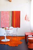 weiße Schalenstühle mit Metallgestell und Klassiker Tisch auf orangem Tierfellimitat, an Wand modernes Bild, seitlich orange Hocker und roter Beistelltisch