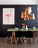 Black classic chairs around table top on wooden trestles, above several copper-colored pendant lights and flamingo picture on black wall