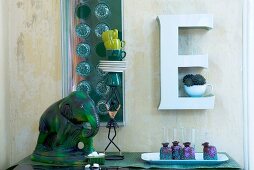Weißer Dekobuchstabe als Wandregal mit grüner Elefantenfigur und kunstvollem Geschirrstapel auf Anrichte