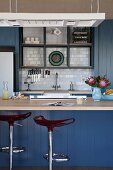 Blue-painted kitchen counter with designer bar stools; white tiled splashback in fitted kitchen in background