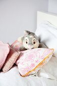 Pale grey rabbit soft toy amongst scatter cushions on bed