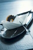 Napkin boat with spoon as rudder and round biscuit as decorative place setting on long, narrow plate