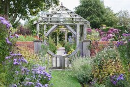 Blooming flower beds in garden with brick wall and wooden gate in front of traditional pergola in silvery wood
