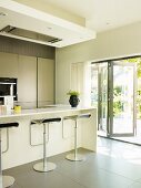 Contemporary, open-plan kitchen with grey tiles floor & bar stools at breakfast bar; open folding doors with view of terrace to one side
