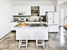 Bar stools at central island with stainless steel worksurface in white, open-plan kitchen