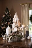 Christmas still-life arrangement on table - dish of fir cones, lit candles and white glass vessel in front of decorated Christmas tree in corner of room