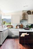 White fitted kitchen with panoramic corner window, wicker lampshade above counter and various cooking utensils arranged on stainless steel worksurface