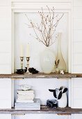 Vases, candlesticks and books on rustic wooden shelves built into niche with white-painted wooden back wall