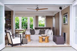 Open-plan living area with sand-coloured walls, pale, comfortable seating, scatter cushions and view of palm trees in summery garden