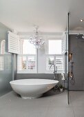 Free-standing designer bathtub in front of open wooden louver shutters and chandelier in grey bathroom with rainfall shower