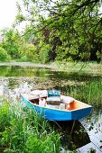 Hellblaues Ruderboot mit Picknick-Utensilien zwischen Schilfpflanzen auf einem Weiher