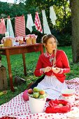 Young woman sitting on picnic blanket peeling apple in front of preserving jars on table and washing line