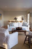 Sofa set with white loose covers and striped scatter cushions around carved wooden coffee table below suspended ceiling with recessed spotlights in minimalist, rustic interior