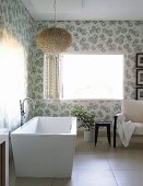 Modern, free-standing bathtub with floor-mounted tape below fluffy pendant lamp in corner of bathroom with floral wallpaper and open window with folding interior shutter