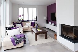 Bright modern interior with white corner sofa, dark wooden coffee table, white rug, purple accent wall and glass-fronted fireplace