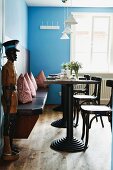 Restaurant with bistro tables, cushions on wooden bench, blue walls and wooden sculpture of man next to open door