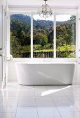 Chandelier and free-standing designer bathtub in sunny bathroom with view over woodland landscape