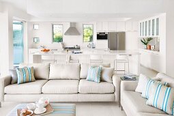 Naturfarbenes Sofa und gepolsterter Couchtisch, im Hintergrund Kochbereich in Weiß mit Theke und Barhockern