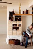 Woman sitting on chair in hall next to chest of drawers, display case and floating shelf