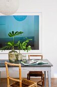 Blau spiegelndes Bild hinter Vintage Tisch mit Metallüberzug und Bambusstühlen; dekorative, grüne Blätter in Glasballons