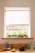 Retro toaster and fruit bowl on kitchen counter below window with grille and half-closed roller blind; flowers in window box outside