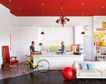Offener Wohnraum mit roter Decke über weißem Küchenbereich und Männer an freistehendem Mittelblock; im Vordergrund roter Ball neben weißem Designer Sofa
