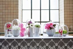 Flower arrangements in white china vases under glass covers on white lace tablecloth in front of industrial window