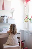 Little girl sitting on stool at white desk and storage boxes on floating shelves