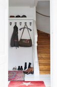 Coat hooks, hat rack and shoes on floor next to foot of staircase in white hall with red rugs