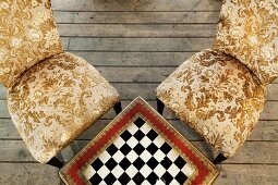 Table with integrated chess board and antique chairs with gold-patterned upholstery on rustic wooden floorboards