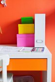 Stack of colourful, cardboard storage boxes and white box file on desk against orange wall