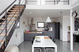 White dining table in front of open-plan kitchen with dark grey cabinets in modern loft apartment with retro ambiance; metal staircase leading to mezzanine