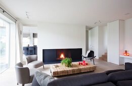 Loungebereich mit Kaminfeuer in modernem Ambiente, graue Sessel und Klassikerstuhl im Retro Look um rustikale Bodentische aus Vierkanthölzern