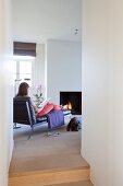 View through narrow doorway of woman sitting on chaise longue in front of fireplace in modern interior