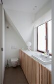 Platzsparendes Bad, vor Fenster eingebautes Waschbecken mit Unterschränken, vertikale Holzlatten Front, im Hintergrund Toilettte