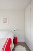 Spherical sconce lamp above red, cylindrical bedside cabinet between bed with red and white blanket and white fitted wardrobes