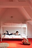 Boy reading on retro-style, white metal bunk beds on orange carpet in converted attic interior with exposed wooden beams