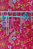 Necklaces of colourful beads hanging from hooks on pink, floral wallpaper