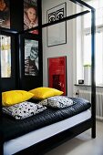 Four-poster bed with black metal frame, yellow and black and white scatter cushions on blanket and collection of posters on wall
