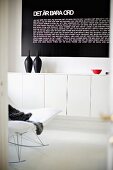 Partial view of modern rocking chair and black vessels on white, floating sideboard below black panel with white lettering