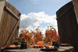 Bundles of straw decorated with flowers on set wooden table in front of open wooden gate with view of cloudy autumn sky