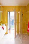 Gelbe Fliesen in Badezimmer mit Waschtisch und bodengleicher Dusche mit Glasabtrennung; Blick durch geöffnete Tür in den Garten