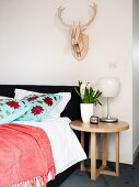 Runder Beistelltisch aus Eichenholz neben Polsterbett mit dekorativ geblümten Kissen und stilisiertes Hirschgeweih an der Wand