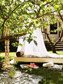 Shady spot in garden - airy canopy above wicker day bed behind picnic blanket and cushions on lawn