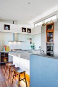 Kitchen island with blue, half-height wall and wooden stools in corner of open-plan kitchen with transom windows