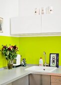 Küchenzeile mit grün lackiertem Spritzschutz an Wand, darüber weisser Hängeschrank; Blumenstrauss, Kaffeemaschine und Schneidebrett mit Kassettenmotiv
