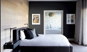 Modern bed with upholstered frame and scatter cushions against matt, varnished wooden wall; wall painted dark grey with doorway leading to ensuite bathroom in background