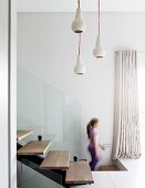 Pendant lamps above modern interior staircase made from steel girders with floating wooden treads and glass balustrade; girl in background